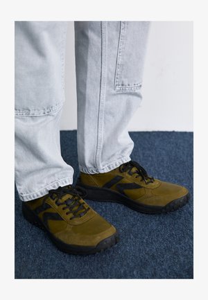 Olivgrüne Sneakers mit schwarzen Akzenten, aus Wildleder und Mesh-Materialien, mit abgerundetem Zehenbereich und strukturierten Sohlen. Kombiniert mit hellgrauen Jeans.