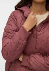 Ženska si prilagaja ovratnik vijoličaste prešite jakne preko krem puloverja, s poudarkom na rebrastih manšetah in detajlih z ježki.