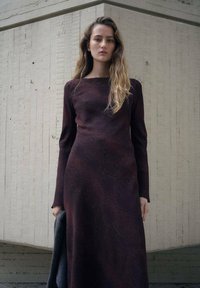 Langarmige, tot op de grond vallende jurk in diep bordeauxrood met subtiele textuur en patroon. Heeft een vierkante halslijn en een getailleerd silhouet.