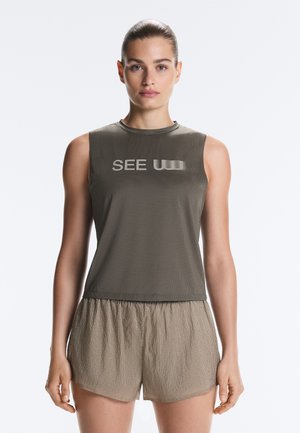 Donna con capelli raccolti, con indosso un top grigio senza maniche con la scritta "SEE" e pantaloncini beige larghi, in piedi davanti a uno sfondo chiaro uniforme.