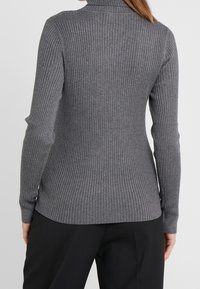 Grijze geribbelde coltrui met lange mouwen, met een aansluitend ontwerp en subtiele textuur. Gezien vanuit de achterkant, gecombineerd met zwarte broek.