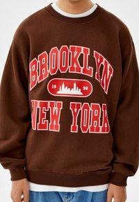 Brun sweatshirt med vit och röd text "BROOKLYN NEW YORK", med en silhuettgrafik och ribbade ärmslut och nederkant. Avslappnad passform.