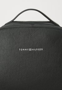 Dos de sac à dos en cuir noir texturé avec poignée cousue et logo Tommy Hilfiger blanc centré sous la poignée.
