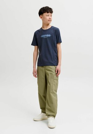 Tienerjongen staat, draagt een marineblauw grafisch t-shirt, olijfgroene cargobroek en witte sneakers, kijkt opzij tegen een effen achtergrond.