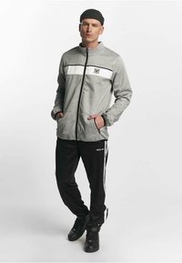 Mann iført grå jakke med glidelås og hvit stripe, svarte joggebukser med hvite side-striper, svarte sneakers og en svart lue, stående med hendene i lommene.