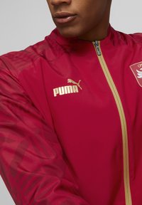 Czerwona lekka kurtka z złotym zamkiem i złotym logo Puma. Przezroczyste wzorzyste rękawy oraz emblemat drużyny na piersi. Gładka tekstura.