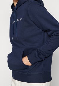 Sudadera azul marino de tela suave, con un bolsillo frontal, puños acanalados y capucha con cordón. Logo "TOMMY JEANS!" en letras estilizadas.