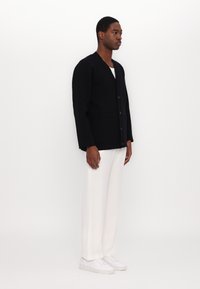 Cardigan noir en tricot avec un col en V, six boutons et deux poches avant. Porté sur un t-shirt blanc et associé à un pantalon blanc et des baskets.