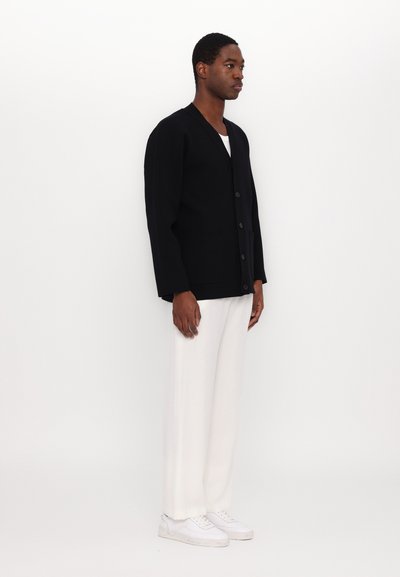 Black knitted cardigan with a V-neck, six buttons, and two front pockets. Worn over a white T-shirt and paired with white trousers and sneakers.