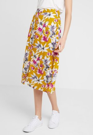 Femme portant une jupe midi jaune à fleurs roses et blanches, associée à un haut blanc et des baskets blanches, debout sur un fond uni.