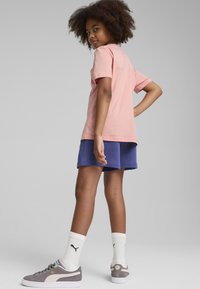 Camiseta de algodón rosa claro, de manga corta, combinada con pantalones cortos morados. Zapatillas grises con detalles blancos y calcetines blancos visibles.