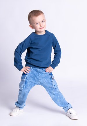 Niebieska bluza z długimi rękawami w połączeniu z jasnoniebieskimi, teksturowanymi spodniami typu cargo z bocznymi kieszeniami oraz białymi sportowymi butami.