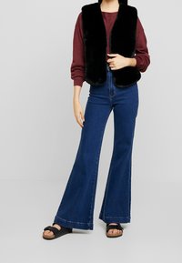 Gilet in pelliccia sintetica nera sopra una maglietta a maniche lunghe bordeaux, abbinato a jeans a vita alta in denim blu a palazzo e sandali neri. Texture liscia, design classico.