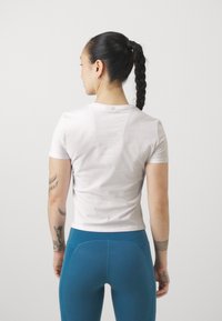 Feszes fehér pamut póló rövid ujjakkal, hátulról nézve, teál leggings-szel párosítva. A pólón kerek nyakkivágás található.