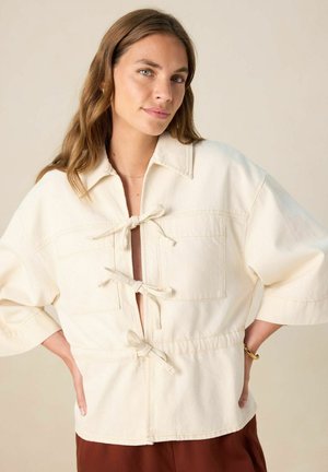 Veste en coton beige avec un col, deux poches avant et des liens décoratifs au niveau du col. Possède des manches amples et une ouverture à l'avant.
