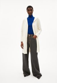 Cremefarbenes, langes Cardigan mit Kapuze und zwei frontalen Taschen, kombiniert mit einem blauen Rollkragenpullover und weiten, dunklen Jeanshosen.