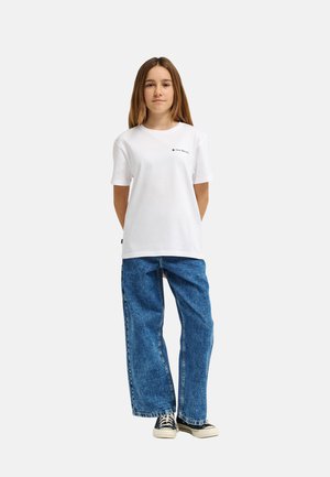 Młoda osoba stoi, nosząc białą koszulkę z krótkim rękawem z małym logo, luźne niebieskie dżinsy i czarne trampki na jednolitym tle.