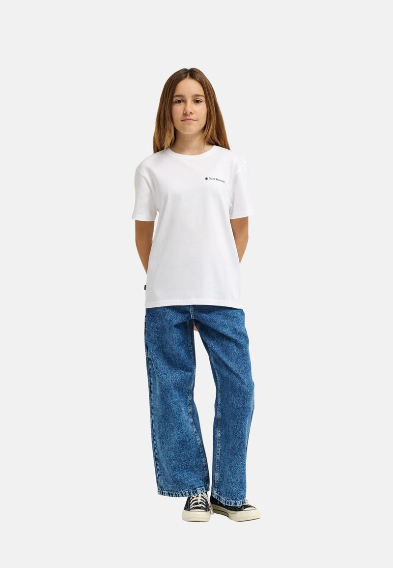 Una persona joven está de pie, vistiendo una camiseta blanca de manga corta con un pequeño logo, jeans azules holgados y zapatillas negras sobre un fondo liso.