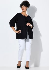 Top nero strutturato con maniche lunghe fino al gomito e dettagli con fiocco, abbinato a pantaloni bianchi con cuciture decorative. Tacchi neri alti.