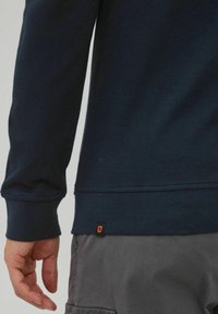 Marineblaues, geripptes Langarmshirt mit abgerundetem Saum, das ein orangefarbenes Markenetikett an der Seite aufweist. Material aus Baumwollmischung. Graue Hose sichtbar.