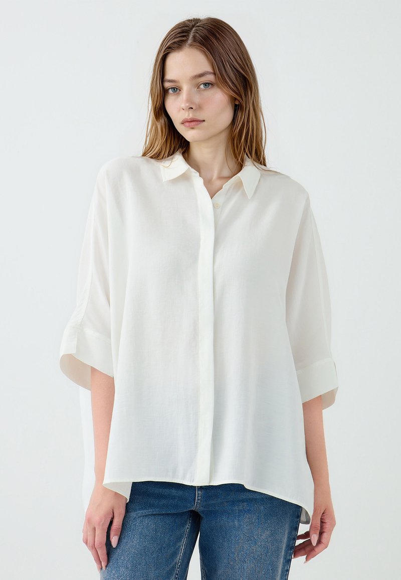 White button-up shirt with short sleeves, made of lightweight fabric. Features a classic collar and a relaxed fit, paired with blue jeans.