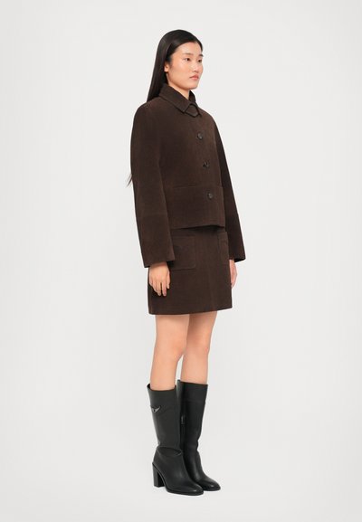 Brown suede cropped jacket with large buttons, paired with a matching mini skirt. Model wears knee-high black heeled boots.