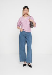 Helles rosa gestricktes Sweatshirt mit V-Ausschnitt, kombiniert mit weit geschnittenen blauen Jeans. Das Outfit wird durch schwarze spitze Schuhe und eine graue Tasche ergänzt.