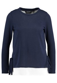 Mörkblå långärmad topp med rund hals, lager av vit fåll och sidoknytning. Mjuk, texturerad tyg med en avslappnad design.