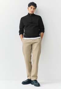 Zwarte ritsvest, beige chino's en zwarte schoenen. Het vest heeft geribbelde manchetten en een hoge kraag. De chino's zijn recht van pijp en hebben een gladde textuur.