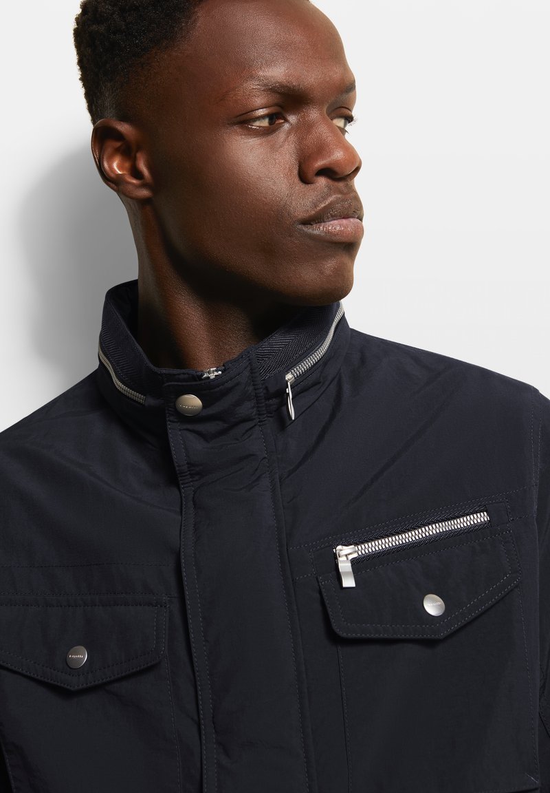 Marineblaue Jacke mit hohem Kragen, Reißverschlusstaschen, silbernen Hardware-Akzenten und einer glatten Textur. Detaillierte Nähte auf der Brust.