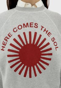 Sweatshirt gris avec un graphisme de soleil rouge audacieux et le texte "ICI VIENT LE SOL" disposé en cercle autour du soleil.