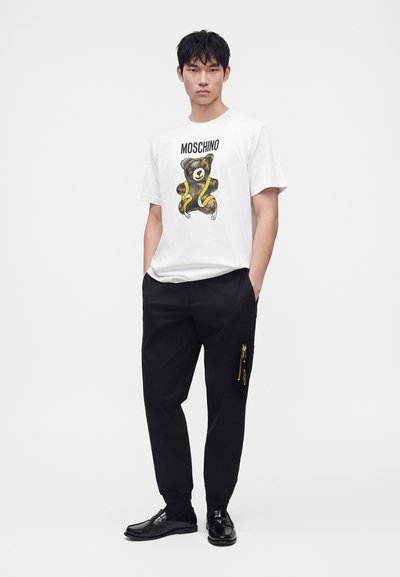 Νέος άνδρας φοράει λευκό μπλουζάκι Moschino με σχέδιο αρκούδας, μαύρο παντελόνι με χρυσά φερμουάρ και μαύρα loafers, στέκεται με τα χέρια στις τσέπες.