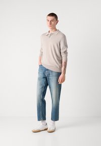 Ljusbeige långärmad polo med ribbade ärmslut, ihopparad med ljusblå tapered jeans och vita sneakers med beige detaljer.