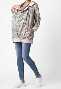 Felpa premaman con motivo floreale, dettagli rosa chiaro, chiusura con zip frontale e tasca a marsupio. Il modello indossa jeans e sneakers bianche.