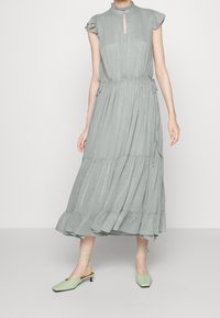 Ärmlös ljusgrå midi-klänning med volangkrage och -kant, samlad midja, kombinerad med ljusgröna låga snörskor.