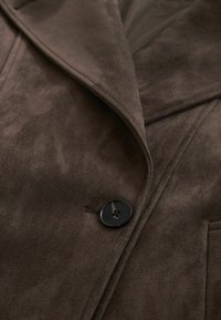 Ruda dirbtinės zomšos paltas su atvartais, vienu juodu sagos užsegimu ir matomomis siūlėmis ant lygaus paviršiaus.