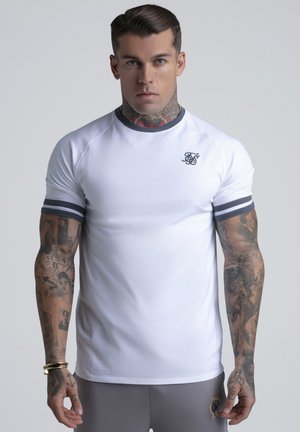 Mann med nakke- og arm-tatoveringer iført hvit tettsittende t-skjorte med mørk kant og logo, stående foran en ensfarget grå bakgrunn.