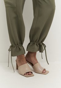 Beige öppna klacktofflor med fyrkantig tådesign. Bärs tillsammans med olivgröna byxor med elastiska muddar och dragsko-detaljer.