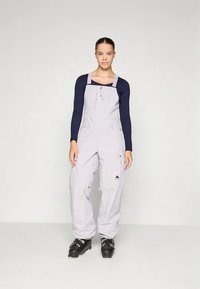 Heledat hallid suusakuubikud reguleeritavate rihmadega, lukuga ja tasku detailidega, kombineeritud tumesinise pika varrukaga särgi ja mustade suusasaabastega.