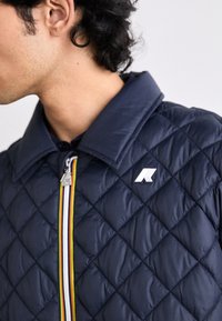 Homme portant une veste matelassée bleu marine avec une fermeture éclair à rayures arc-en-ciel et un logo blanc sur la poitrine, vu de profil devant un fond uni.