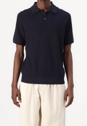 Mand iført en marineblå kortærmet strikket poloshirt kombineret med løstsiddende, off-white bukser mod en ensfarvet baggrund.