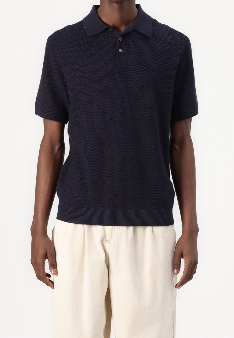 Homme portant un polo en maille bleu marine à manches courtes, associé à un pantalon ample couleur écru, sur un fond uni.