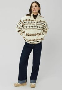 Pull en polaire crème avec des motifs géométriques marron, col zippé mi-haut et poche avant, associé à un jean large bleu foncé et des chaussures crèmes.
