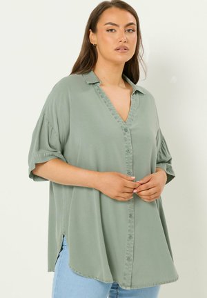 Frau trägt lockeres, salbeigrünes Hemd mit aufgekrempelten kurzen Ärmeln und hellblaue Jeans, steht vor neutralem Hintergrund.