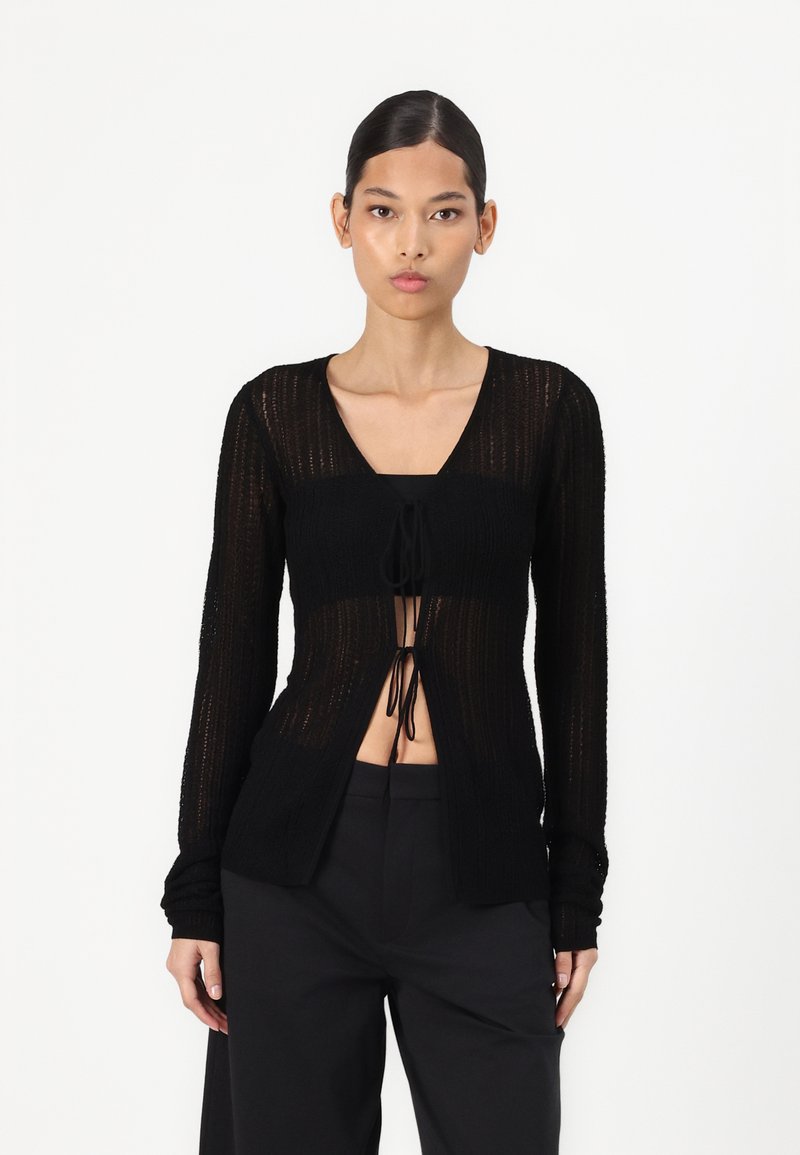 Cardigan nero a maglia con maniche lunghe, apertura frontale e chiusura con coulisse. Motivo testurizzato e silhouette aderente. Abbinato a pantaloni neri.