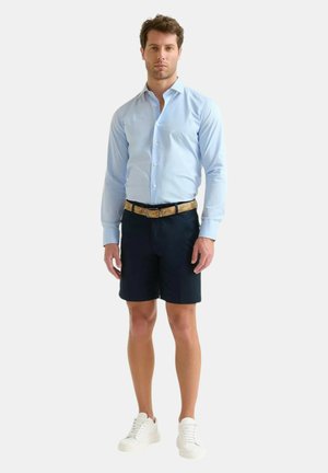 Camicia azzurra chiara con bottoni, pantaloni corti scuri, cintura beige con motivo, sneaker bianche, postura in piedi, sfondo neutro.