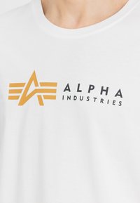 Balta medvilninė marškinėliai su juodu ir oranžiniu logotipu, kuriame yra abstrakti žvaigždės forma ir užrašas "ALPHA INDUSTRIES" paryškintomis raidėmis.