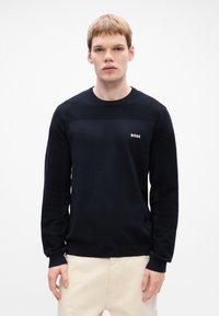 Tânăr îmbrăcat cu un pulover BOSS negru și pantaloni crem, stând în fața unui fundal alb simplu, cu fața spre cameră și o expresie neutră.
