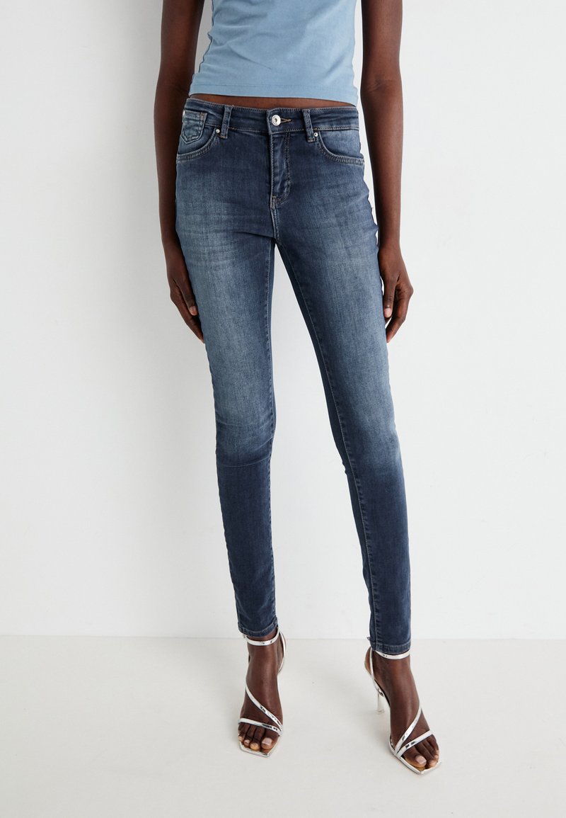 Jeans attillati in denim blu scuro con lavaggio sbiadito, dotati di tasche frontali e chiusura con bottone. Indossati con tacchi alti argentati con cinturini.