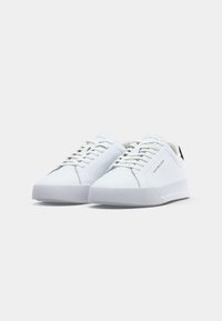 Paire de baskets basses blanches avec lacets, semelles fines et marquage "Tommy Hilfiger" sur le côté et la languette, montrée sur un fond uni.